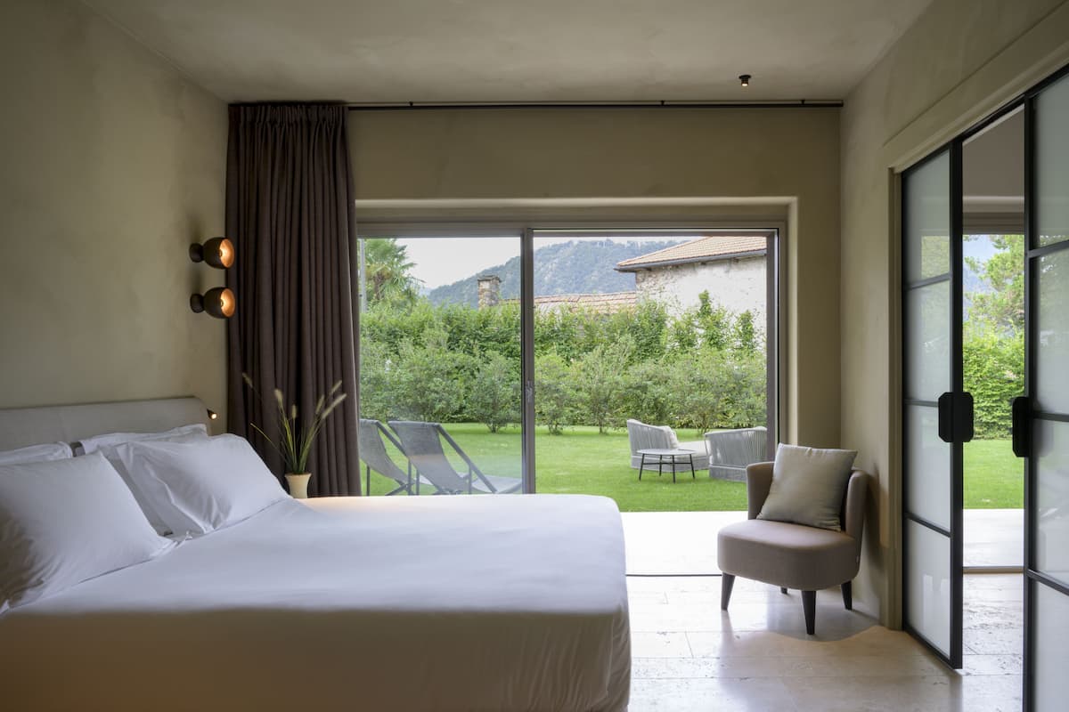 Een slaapkamer met een groot bed, een stoel en uitzicht op een tuin en bergen door een glazen schuifdeur.