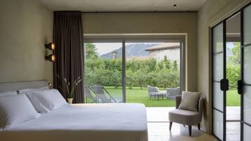 Un dormitorio con una cama grande, una silla y vistas a un jardín y montañas a través de un ventanal corredero.