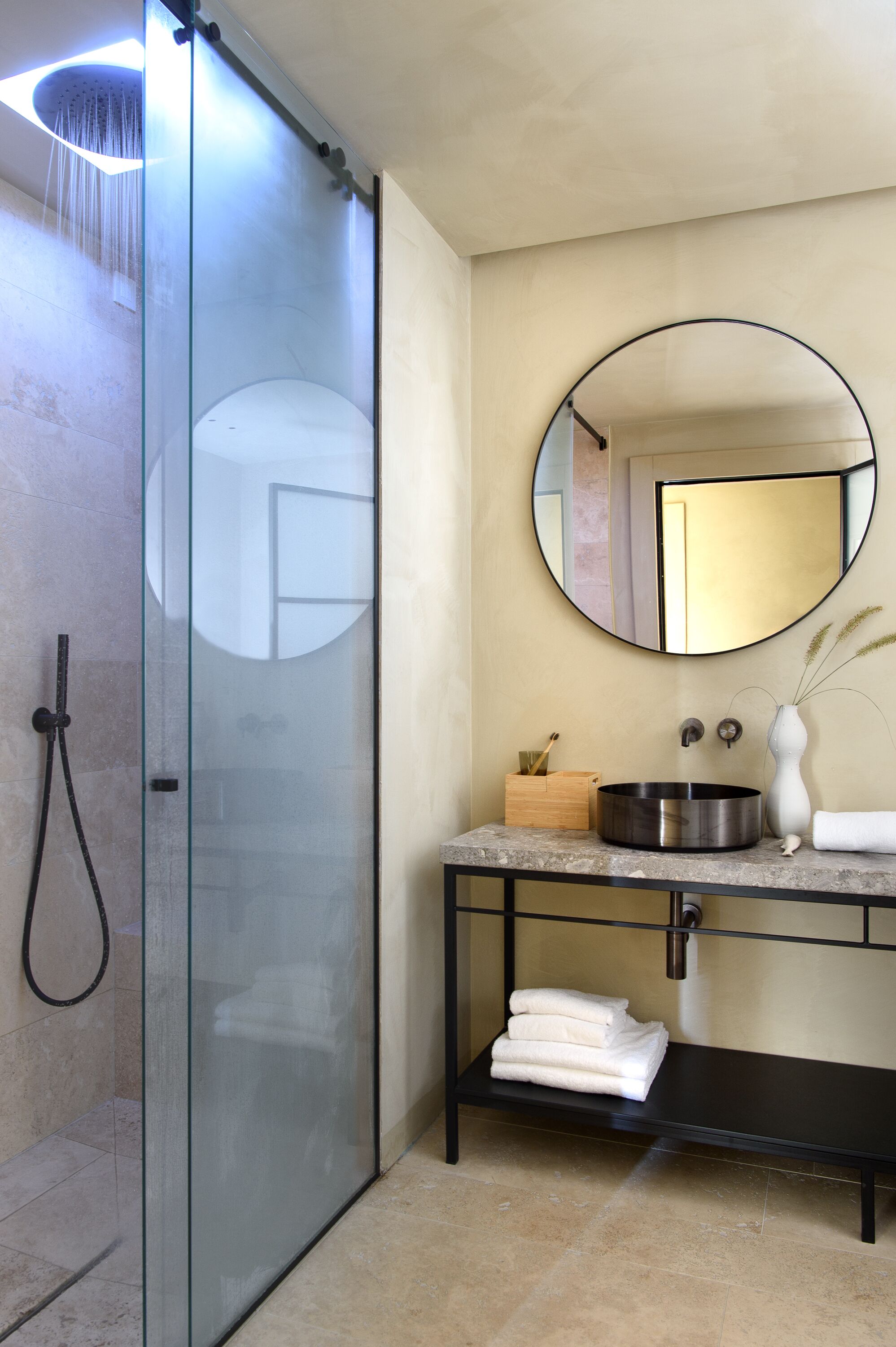 A modern bathroom with a glass-enclosed shower, a round mirror, a sink with a vase, and neatly folded towels on a shelf.