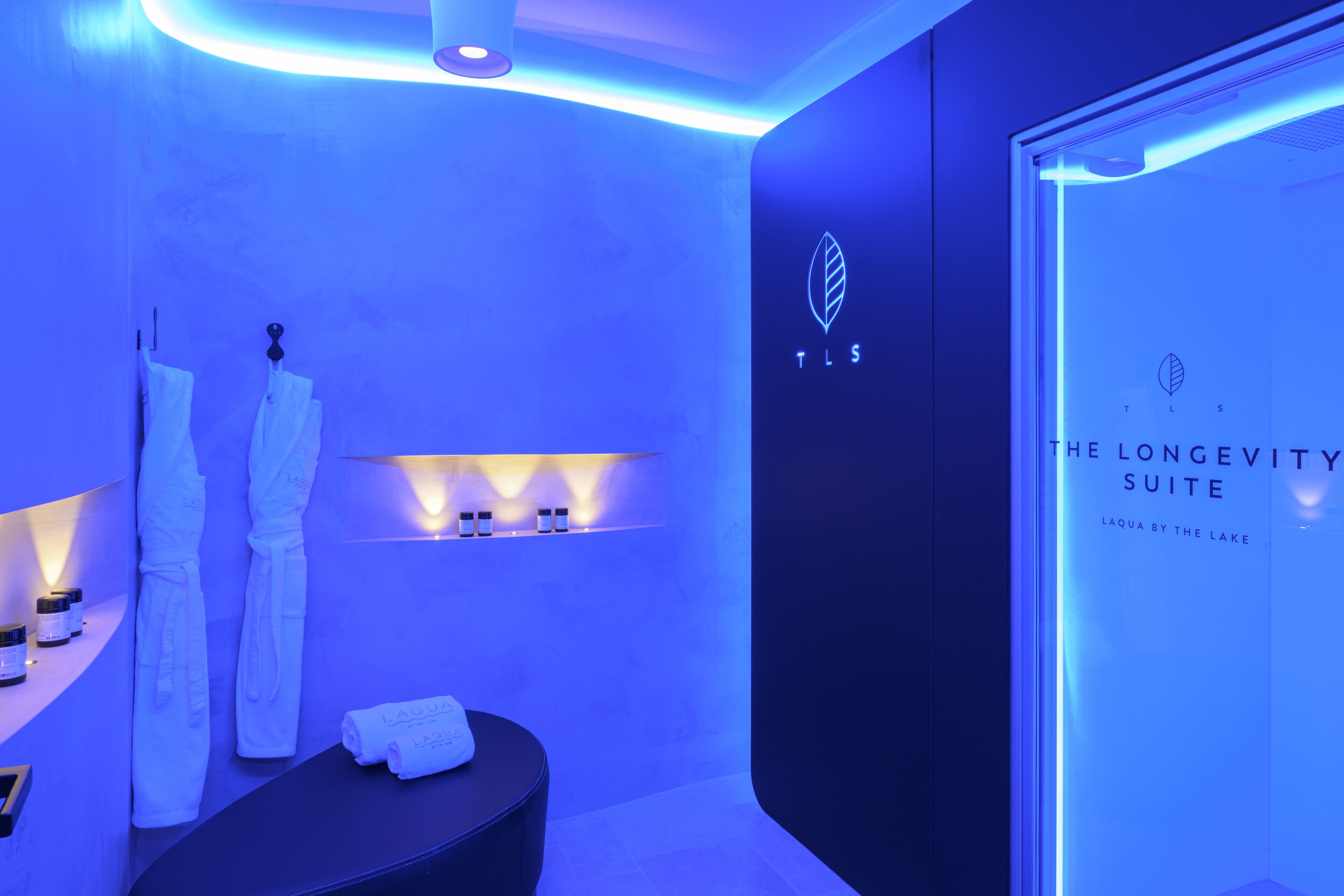 A modern bathroom with a blue ambient lighting, a towel rack, and a glass door with the text "TLS THE LONGEVITY SUITE".