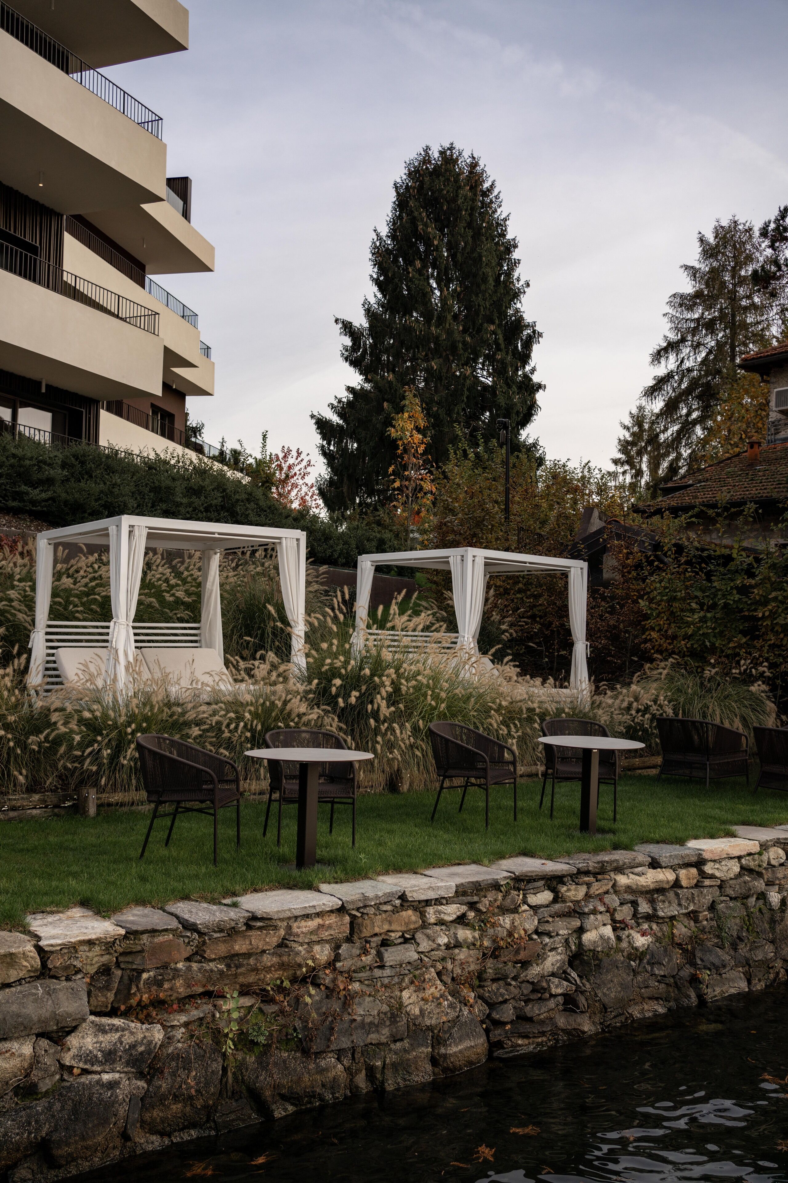 A serene outdoor seating area with white canopied structures, stone walls, and a calm water body.
