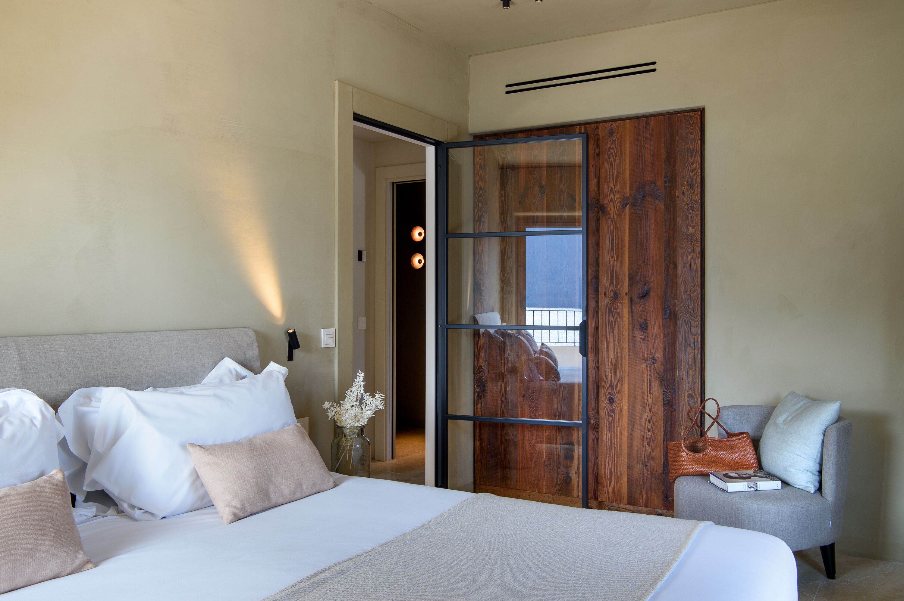 A bedroom with a bed, a grey armchair, a wooden door, and a view through the door.