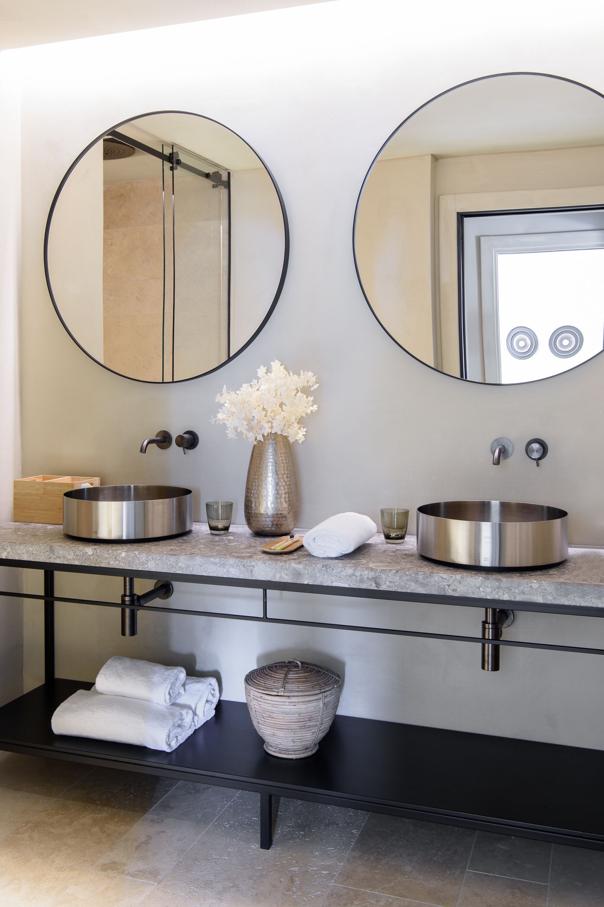Un baño moderno con dos espejos redondos, un lavamanos de acero inoxidable y un florero con flores blancas.