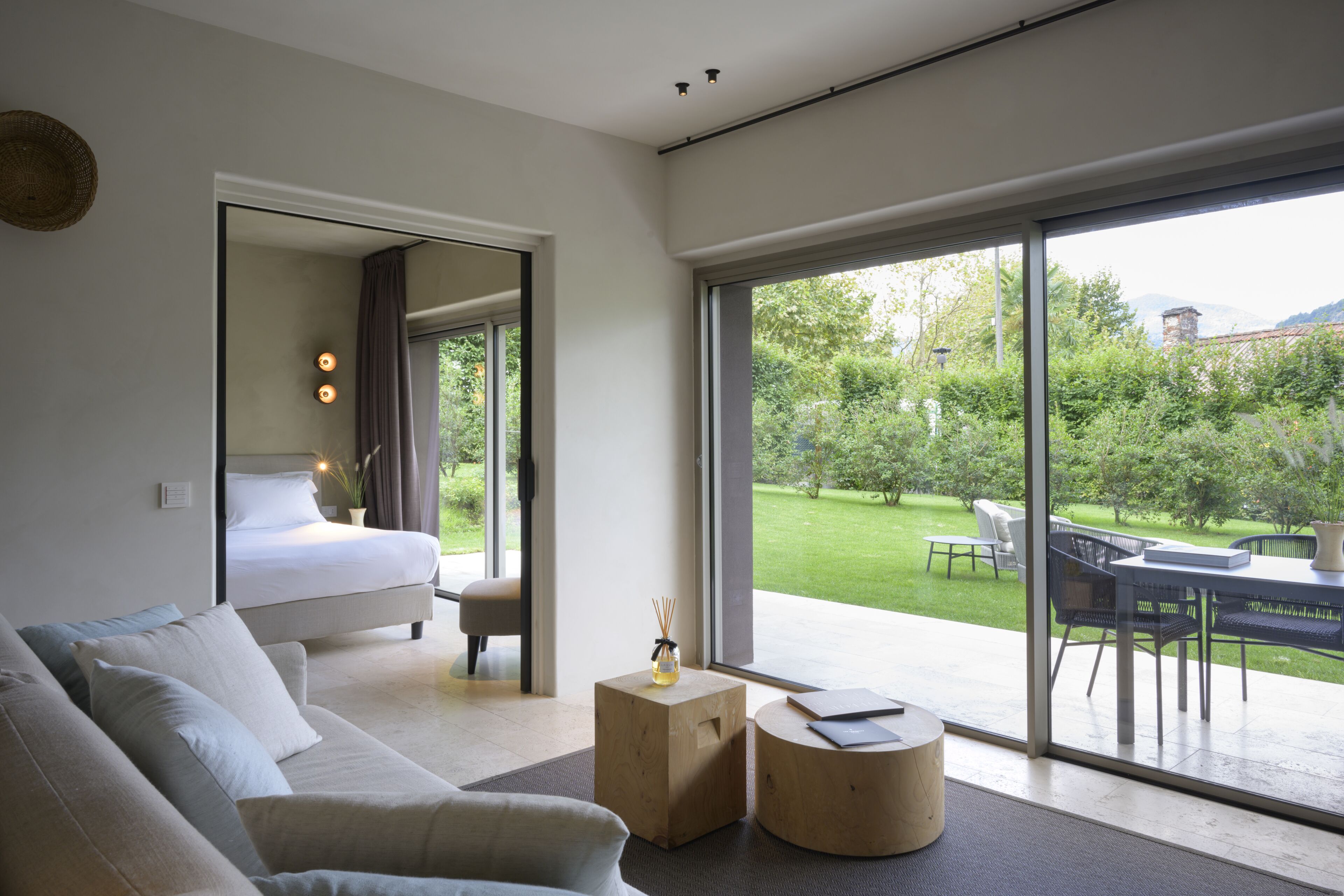 A modern bedroom with a large bed, a sofa, and a view of a garden through a sliding glass door.