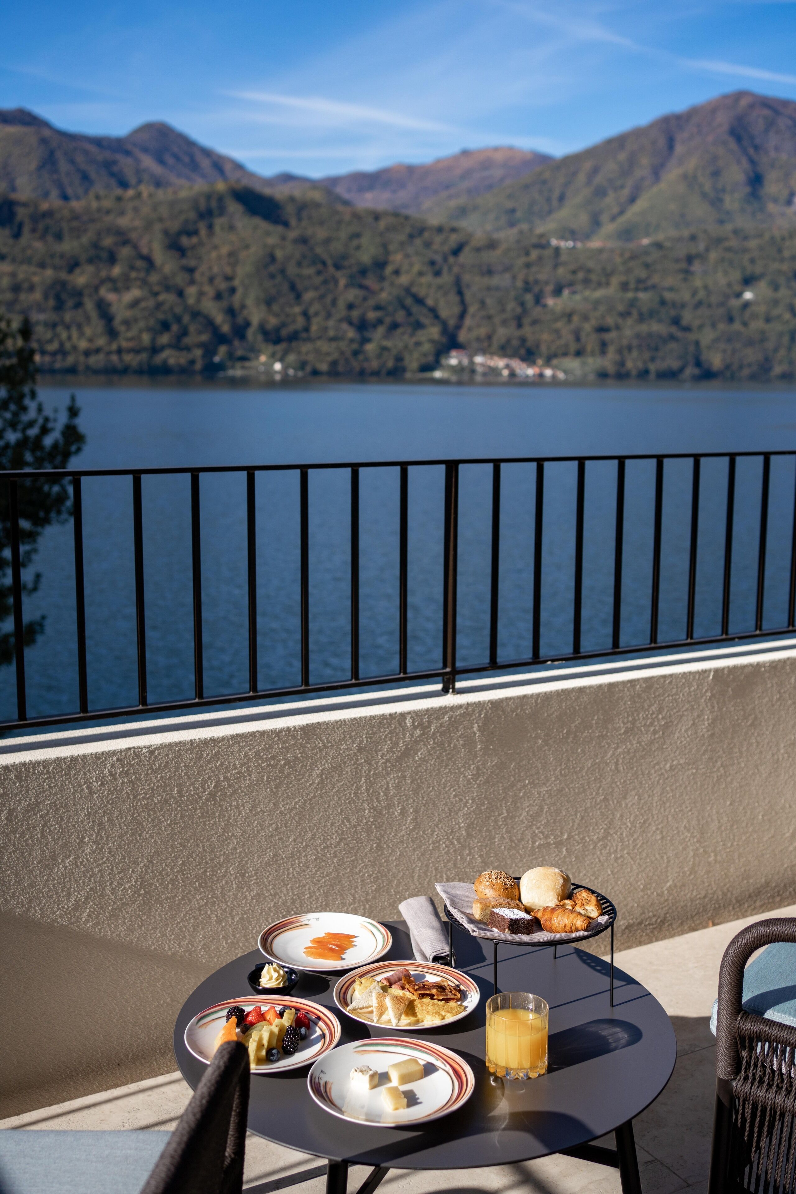 Una mesa con alimentos para el desayuno y un vaso de jugo de naranja en un balcón con vista a un lago y montañas al fondo.