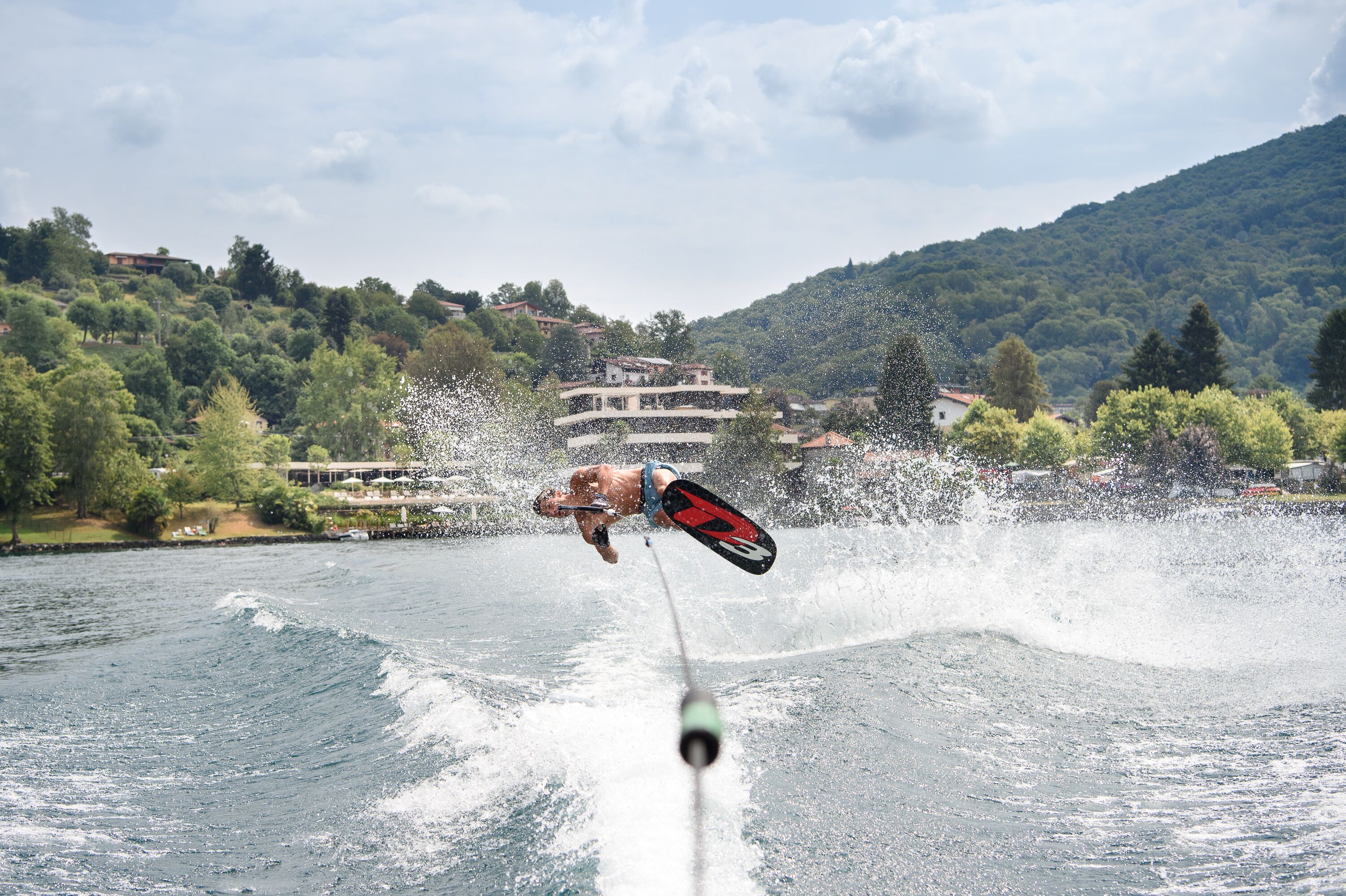 A water sport activity with a wakeboarder in mid-air, a boat, and a mountainous landscape with buildings in the background.