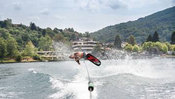 A water sport activity with a wakeboarder in mid-air, a boat, and a mountainous landscape with buildings in the background.