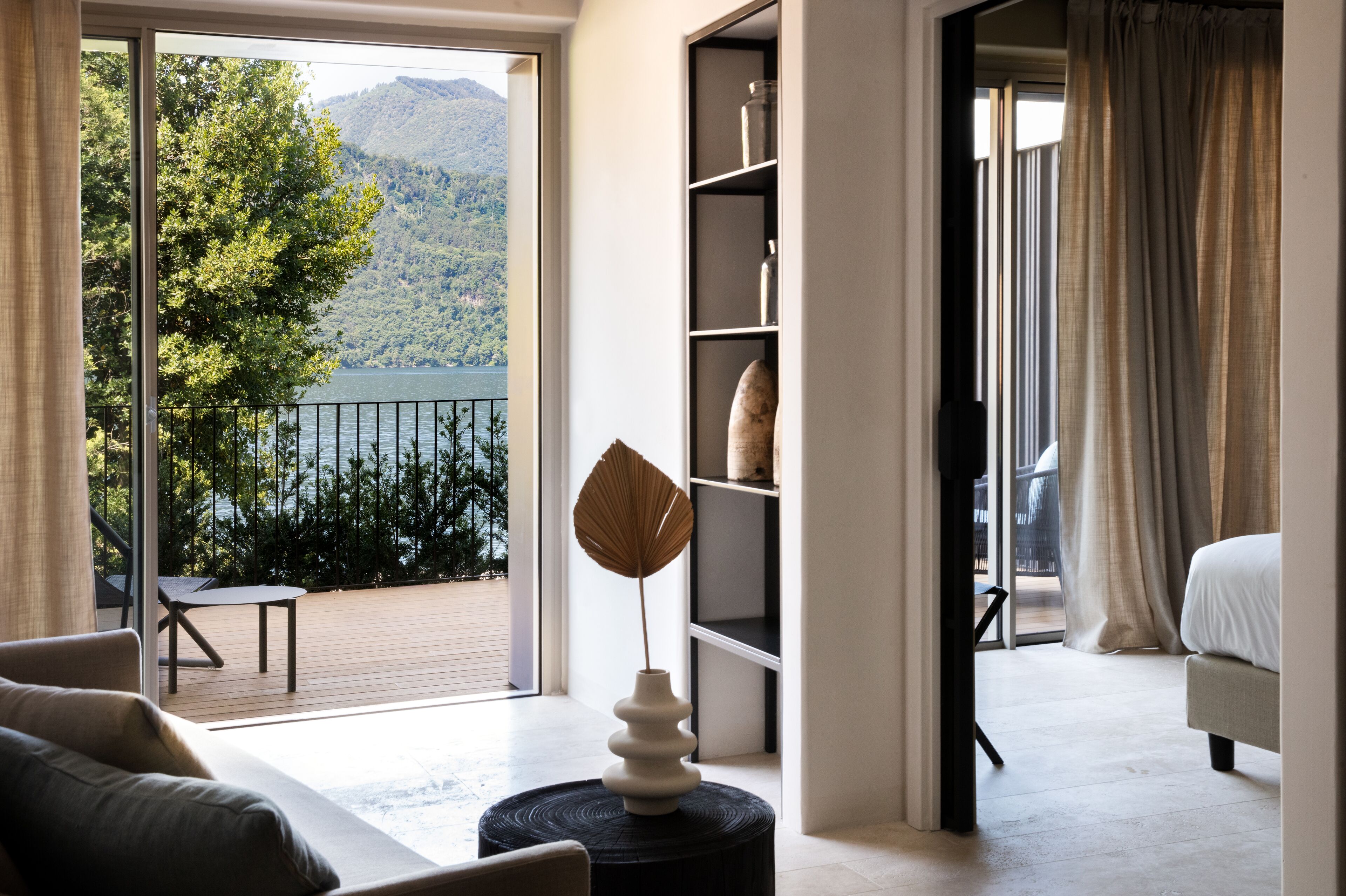 Ein modernes Hotelzimmer mit Balkon, Blick auf einen See und Berge, einem großen Fenster und einem Bett.