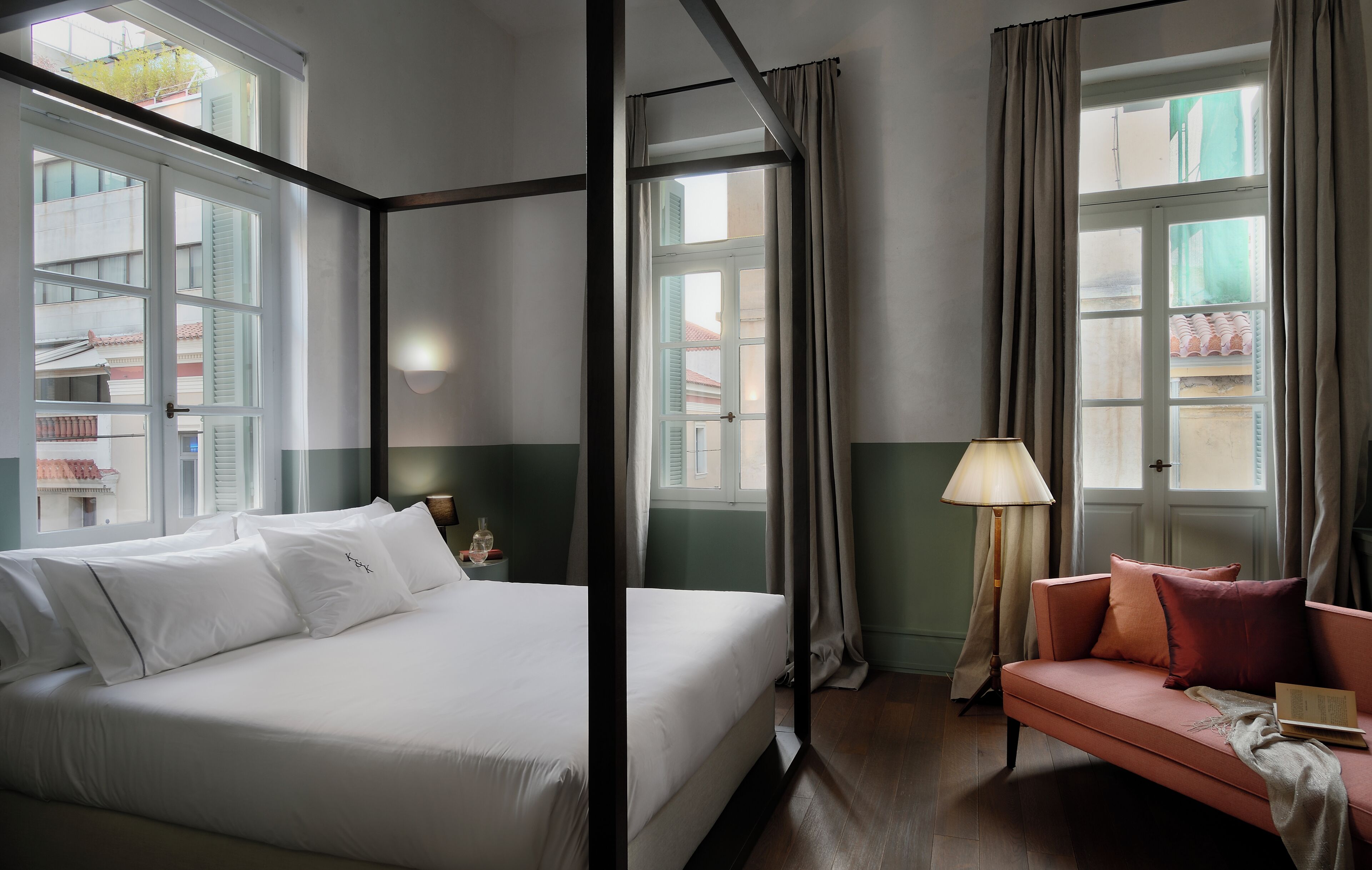 Ein Hotelzimmer mit einem großen Bett, einer Couch, einer Lampe und Blick auf Gebäude durch die Fenster.