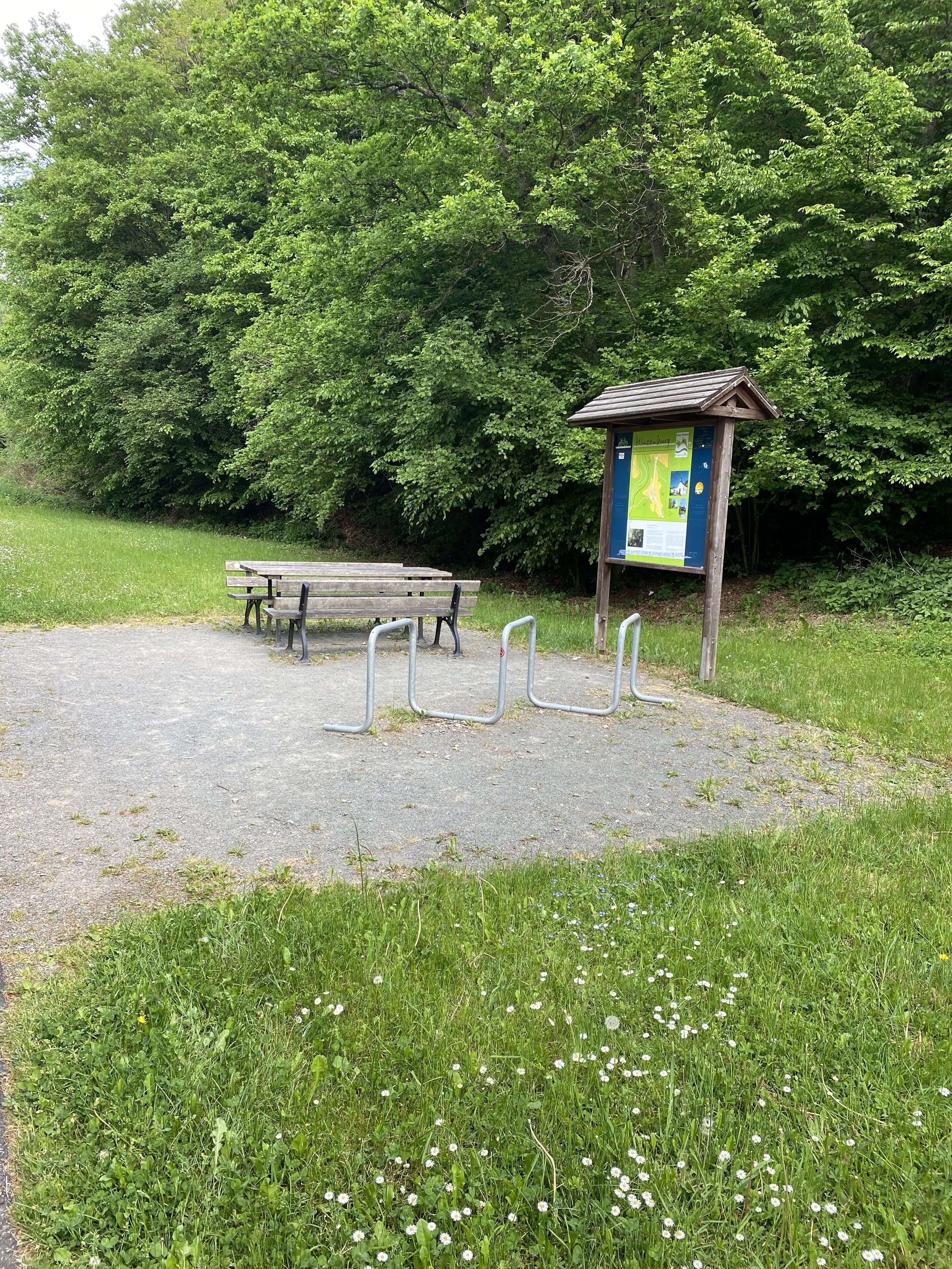 Un parc avec un banc en bois, un panneau d’information en métal et une zone pavée entourée de pelouse et d’arbres.