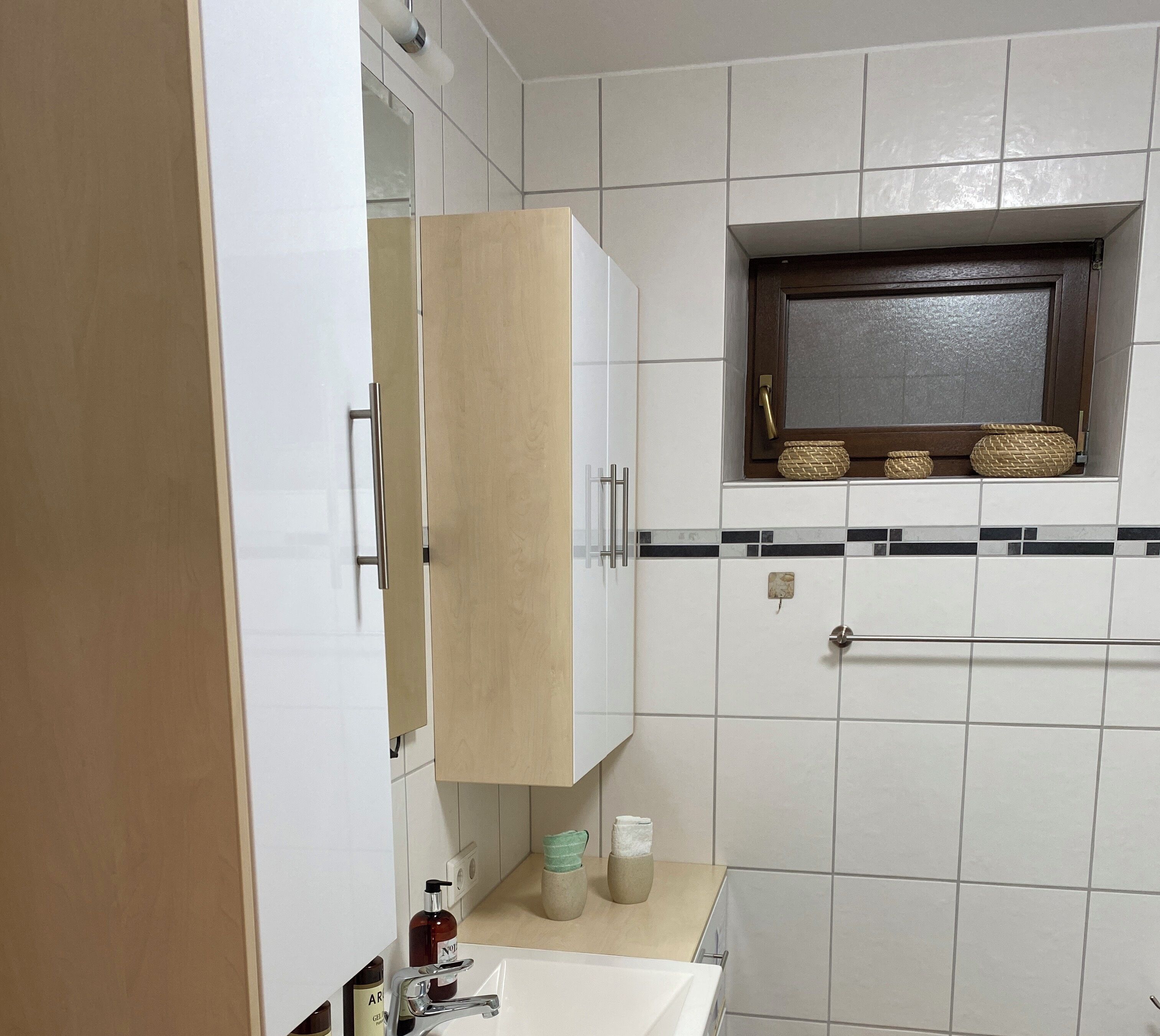 Un baño con un lavamanos blanco, un espejo sobre él y una ventana con cestas tejidas.