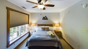 Quarto com cama, duas mesas de cabeceira com luminárias, um ventilador de teto, uma janela com persianas e um objeto decorativo na parede.