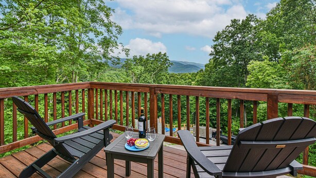 Una terraza de madera con dos sillas Adirondack, una mesa con una botella de vino y un plato de uvas.