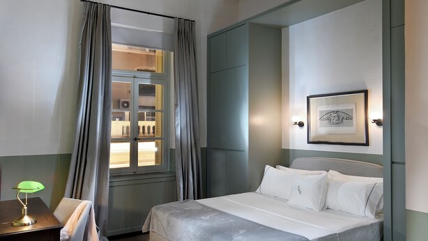 Ein Hotelzimmer mit einem Bett, einem Schreibtisch mit Lampe, einem Sessel, einem Fenster mit Vorhängen und einem Bild in einem Rahmen an der Wand.