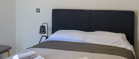 A neatly made bed with a grey quilt and white pillows, a bedside lamp, and a wall-mounted air conditioner.