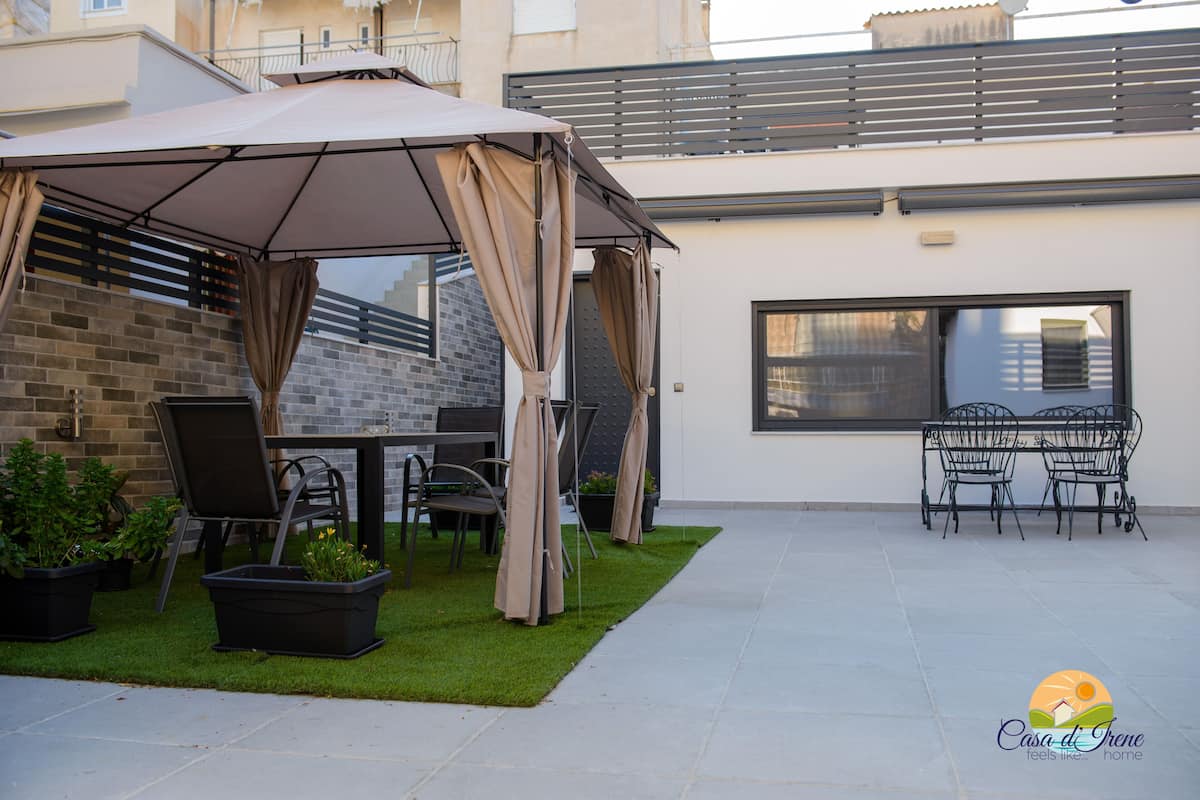 Un espace extérieur aménagé en patio, avec un coin salon couvert, une table et des chaises. On y trouve également des plantes en pot et une petite zone recouverte de gazon artificiel.