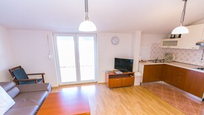 A living room with a sofa, a wooden coffee table, and a television. There is a kitchen area with wooden cabinets and a microwave.