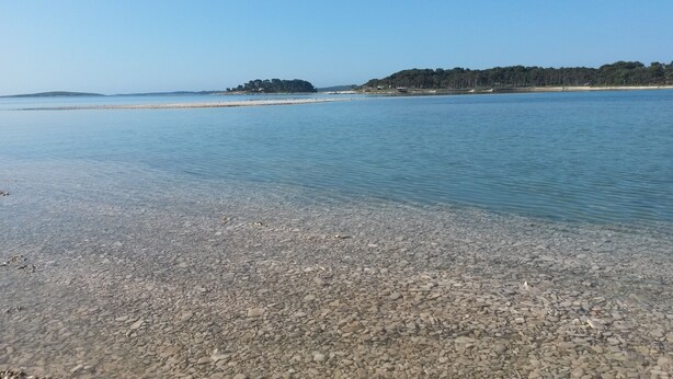 A pebbled beach with clear water, a distant shoreline, and a forested hill.