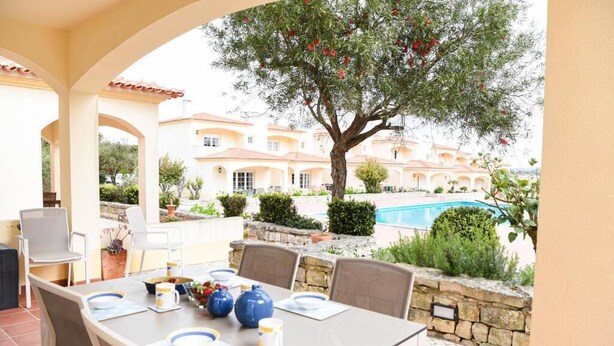 Un área de comedor al aire libre con una mesa dispuesta para cuatro personas, rodeada de sillas, un muro de piedra y una piscina al fondo.
