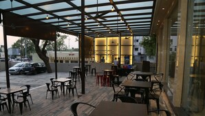 An outdoor cafe with tables and chairs, a covered area with a ceiling fan, and a view of parked cars and buildings in the background. - Hotel Ruturaj Regency (Dhule)
