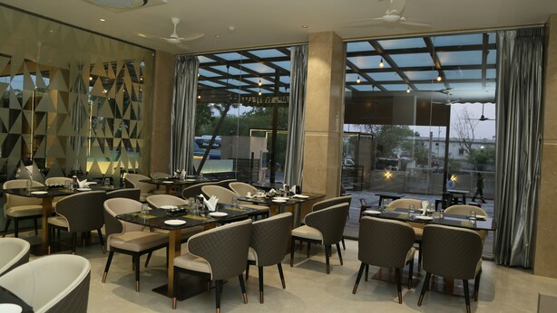 A modern dining area with multiple tables and chairs, large windows with curtains, and a ceiling fan. - Hotel Ruturaj Regency (Dhule)