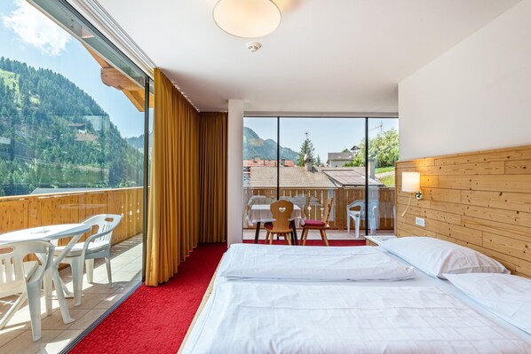 A hotel room with a bed, a small table, chairs, a balcony with a view of mountains, and a wooden headboard.