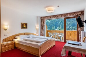 A bedroom with a wooden bed, bedside tables, a desk, a chair, a TV, and a balcony with a view of trees.
