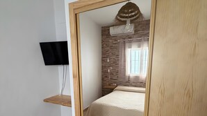 A bedroom with a wooden wardrobe, a bed with a beige bedspread, a mounted TV, and a window with blinds.