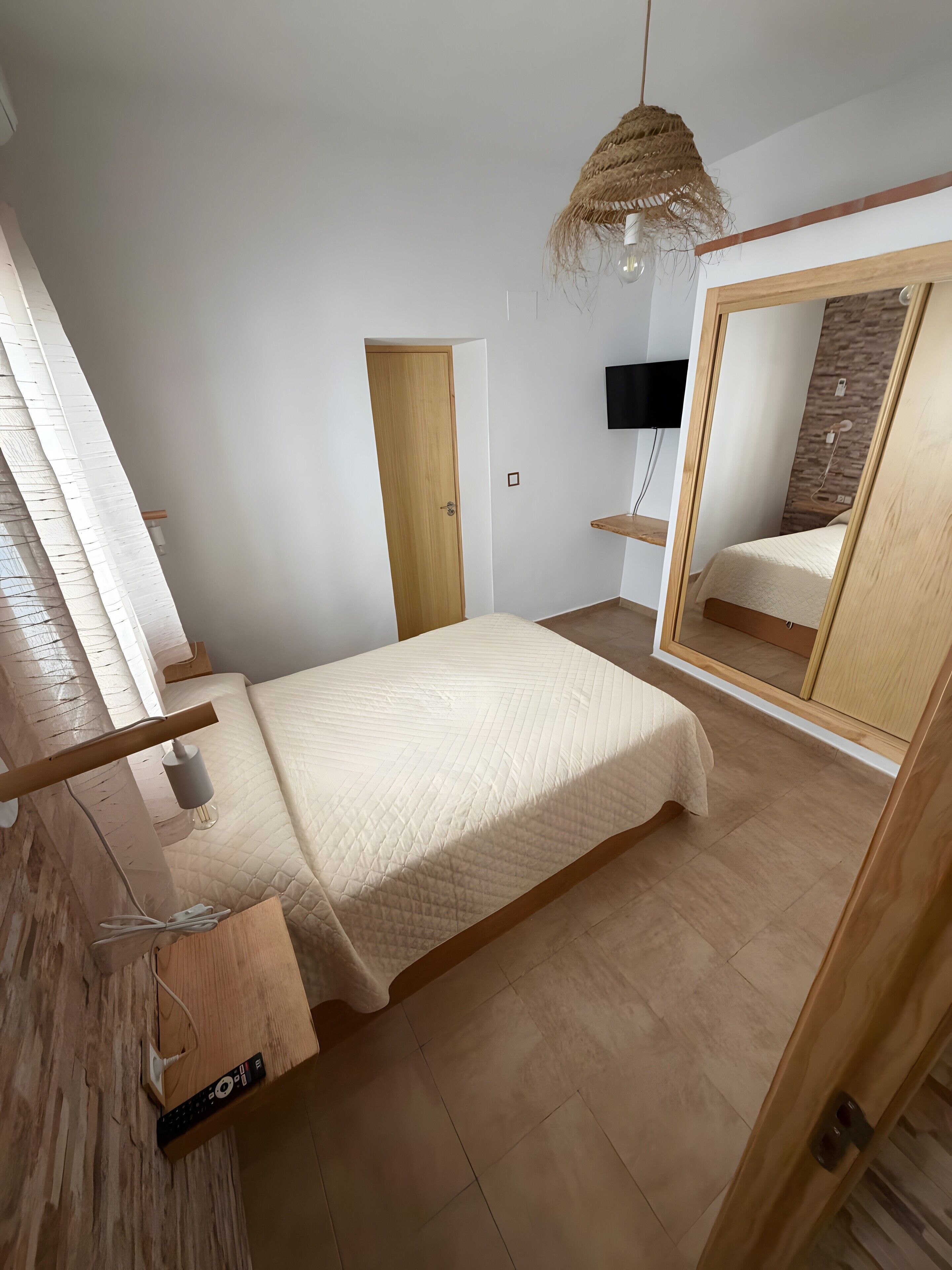 A bedroom with a bed, a wooden floor, a door, a wall-mounted TV, and a mirror.