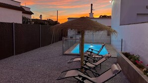 A pool area with lounge chairs, a thatched roof structure, and a clear sky.