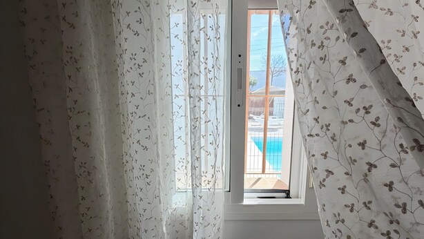 A bedroom with a window, sheer curtains, and a view of a building and a pool.