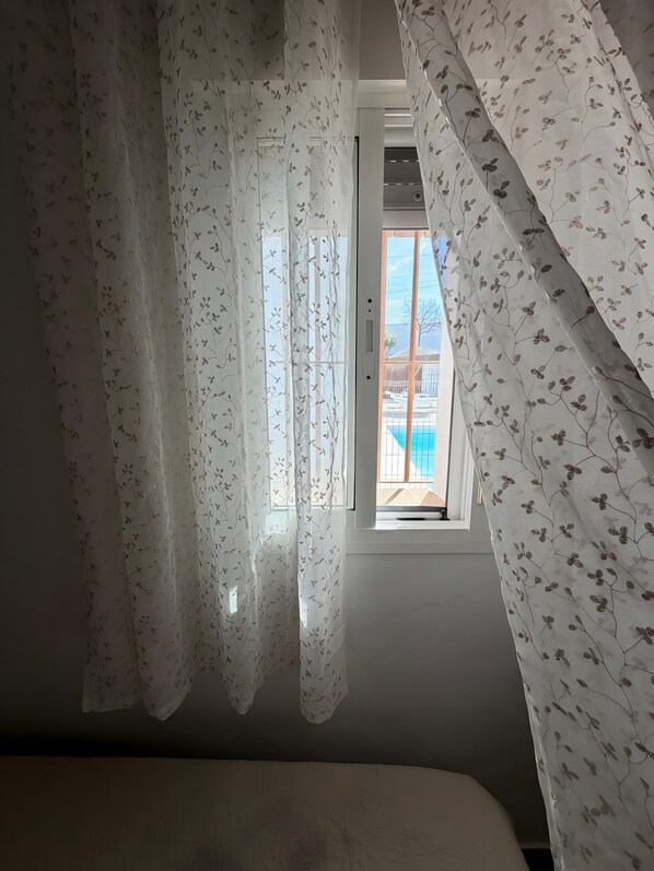 A bedroom with a window, sheer curtains, and a view of a building and a pool.