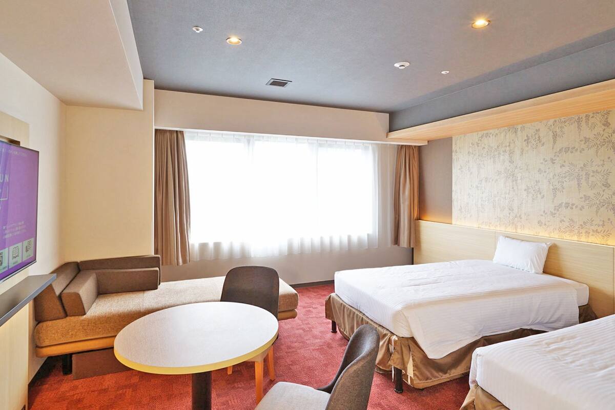Une chambre d’hôtel avec deux lits, une télévision, un canapé, une petite table et une fenêtre avec des rideaux.