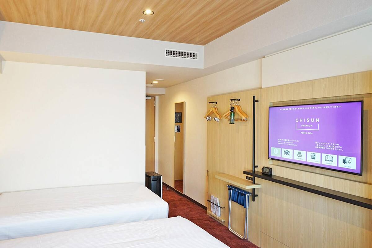 Une chambre d’hôtel avec deux lits, un plafond en bois, un téléviseur fixé au mur diffusant l’émission « CHISUN PREMIUM Kyoto Kyo », et un mur en bois muni d’un porte-serviettes.