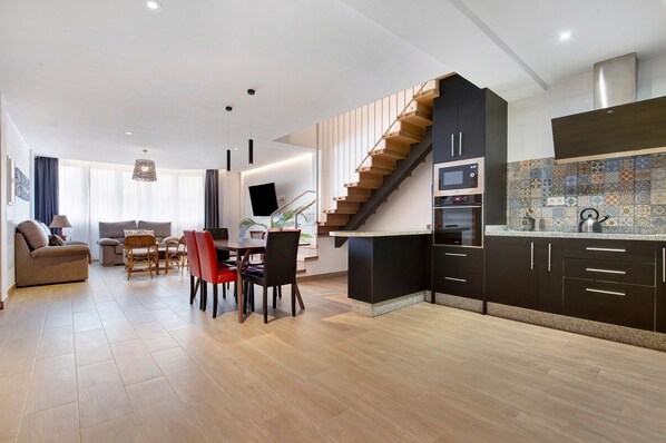 A modern kitchen with a wooden staircase, a dining area with a table and chairs, and a living room with a sofa and a TV.