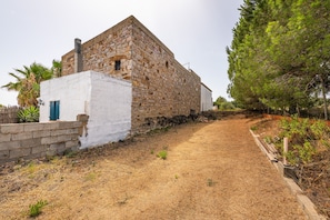 Un edificio de piedra con fachada blanca, un estrecho sendero de tierra y un árbol alto.