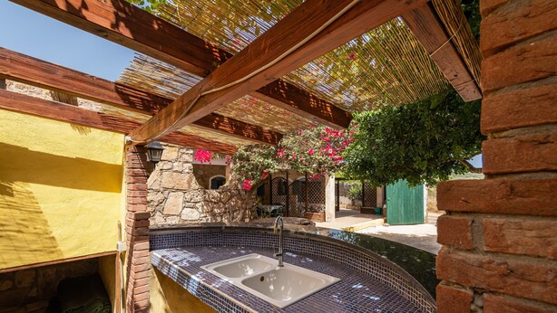 Un espace extérieur abrité, doté d’un évier aux carreaux bleus, de murs en pierre et d’une pergola en bois.