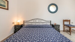 Une chambre à coucher avec un lit, une table de chevet avec une lampe, une chaise en bois, un miroir rond et un tableau encadré accroché au mur.