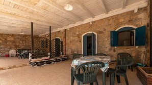 Une pièce aux murs de pierre, dotée d’un plafond haut, d’une table entourée de chaises et d’une fenêtre aux volets bleus.