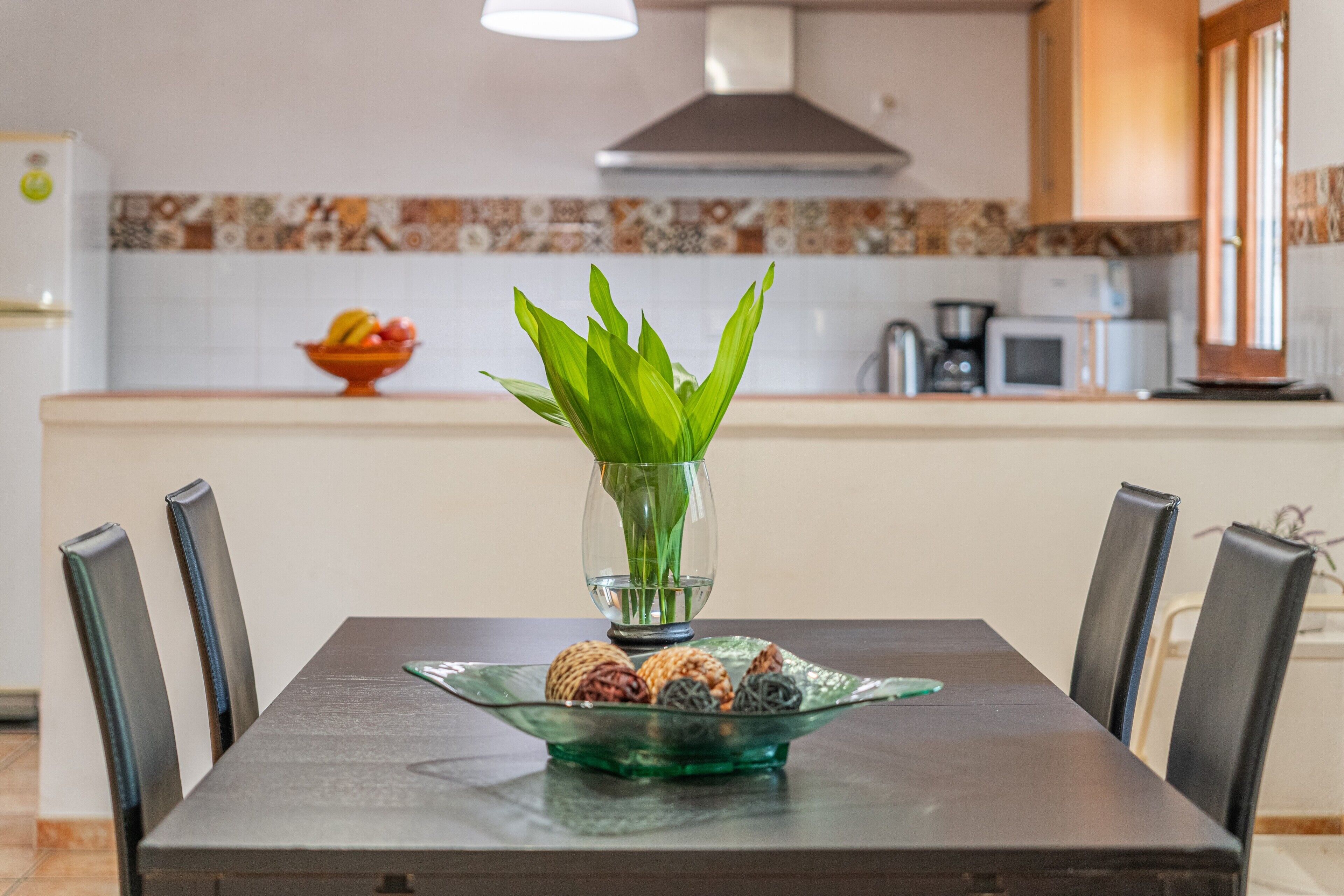 Una mesa de comedor con sillas, un jarrón con hojas verdes y un bol con aperitivos.