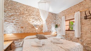 A bedroom with a stone wall, a canopy bed, a wooden headboard, and a window with curtains. - Holiday Home in Es Cami Vell with Pool and Wi-Fi (INCA)