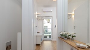 Un couloir étroit doté d’un plancher en bois, de murs blancs, d’une porte en verre donnant sur un balcon, et d’un comptoir en bois orné de plantes en pot et d’un bol.