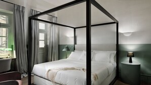 Un letto a baldacchino con lenzuola bianche, un comodino con una lampada e vista sulla città attraverso una finestra con tende.