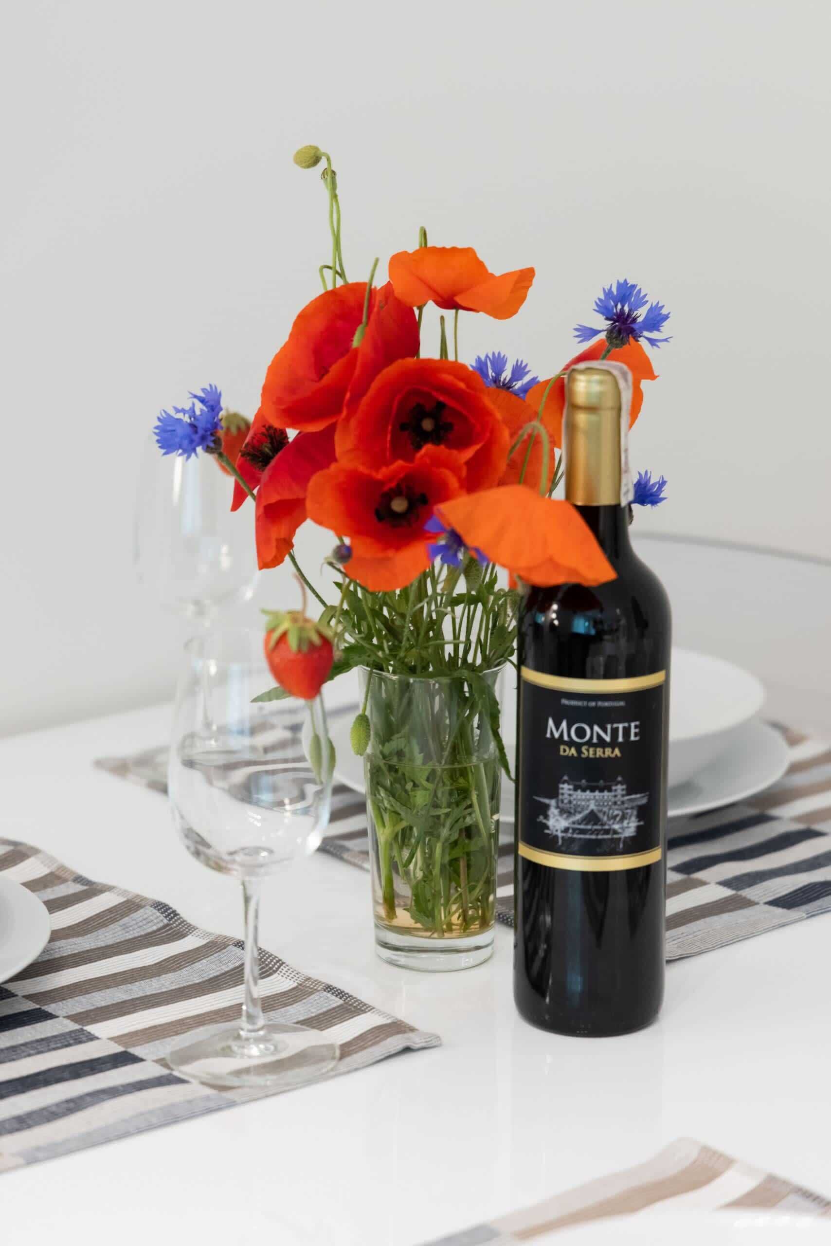 Una botella de vino Monte Da Serra, un jarrón de cristal con flores rojas y otro jarrón de cristal con tallos verdes.