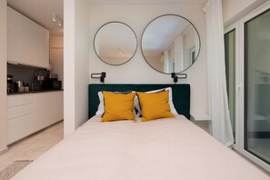 Une chambre à coucher avec un lit, deux miroirs placés de chaque côté du lit, une tête de lit verte et une vue sur une cuisine et un balcon.