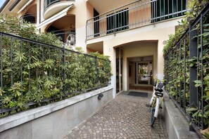 A narrow cobblestone pathway leading to a building entrance, flanked by lush green plants and a bicycle parked by the entrance.