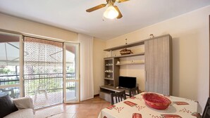 A room with a sliding glass door to a balcony, a dining table with a tablecloth, a television, and a cabinet with shelves.