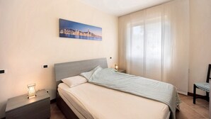 A bedroom with a bed, nightstand, lamp, chair, and a cityscape painting on the wall.
