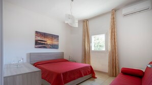 Ett sovrum med en säng, en röd soffa, ett sängbord, ett glas, en tavla och ett fönster med gardiner.