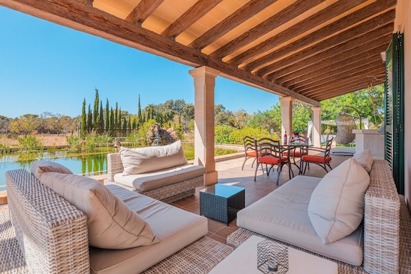 Un patio exterior con mobiliario de mimbre, una mesa de piedra y vistas a un lago y árboles.